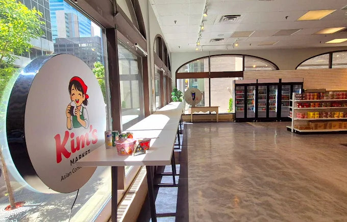 Inside Kim's Market, the round backlit shop sign glowing beside the window onto 5th Avenue